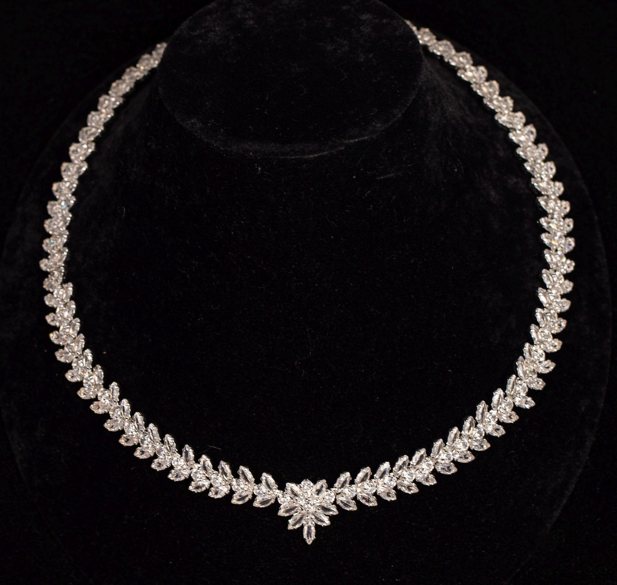 Silver-tone zircon necklace with a continuous row of leaf motifs and a central flower detail on a black velvet bust.