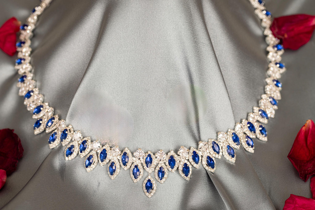 Blue zircon halo necklace on gray satin, showing its graduated stone arrangement and central drop beside red rose petals.