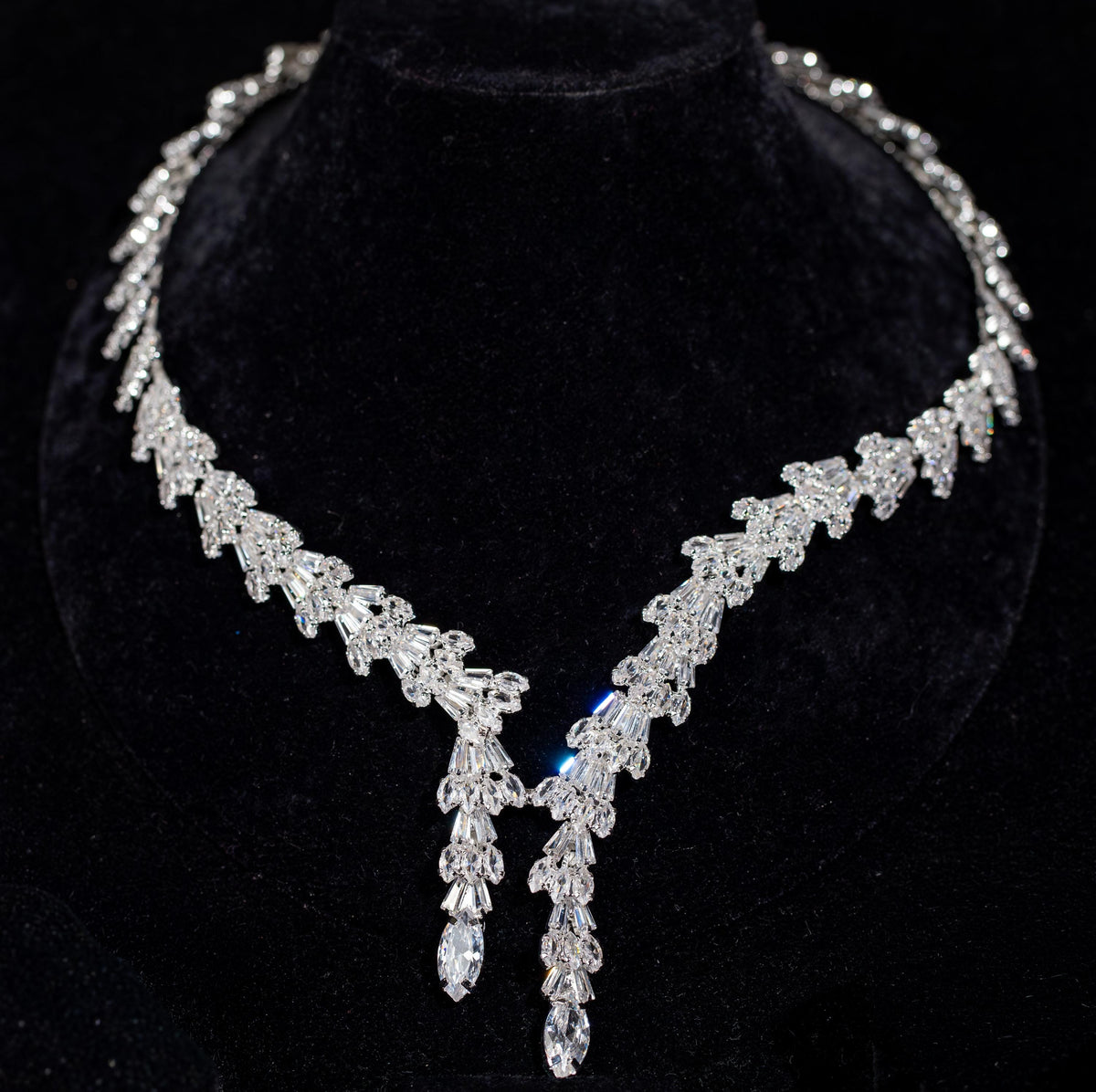 Front view of a baguette zircon necklace with repeating fan-shaped motifs and two pear-tipped drops on a black velvet bust.