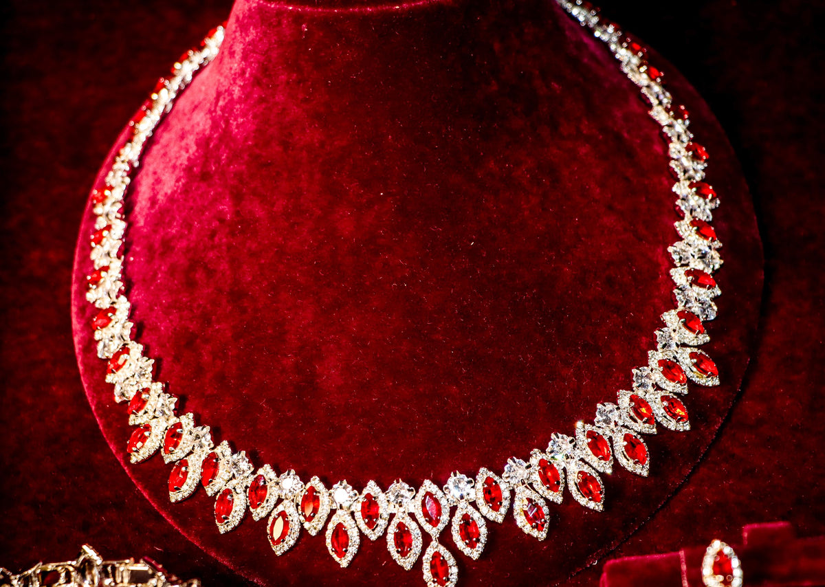 Close-up of a silver-tone necklace with oval red zircon stones framed by clear halos on burgundy velvet.