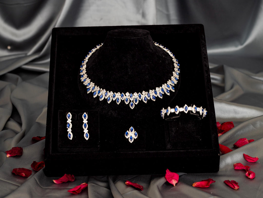 Four-piece blue zircon jewelry set in a black velvet box surrounded by gray satin and scattered red rose petals.